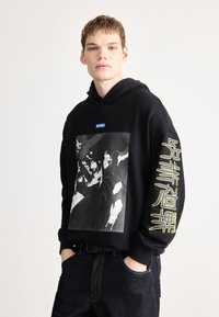 Μαύρο φούτερ με κουκούλα που διαθέτει εκτύπωση γραφικού, ριμπ μανσέτες και μπλε λογότυπο HUGO στο στήθος. Κίτρινα κείμενα στη μανσέτα προσθέτουν αντίθεση.