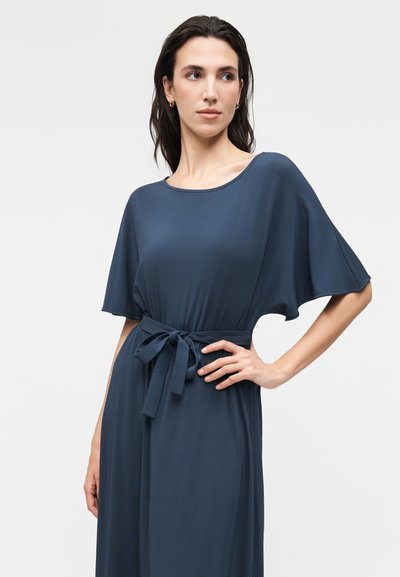 Femme portant une robe bleu foncé avec de larges manches courtes et une ceinture nouée à la taille, regardant légèrement vers sa gauche sur un fond uni.