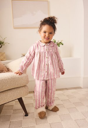 Malé dítě v růžových pruhovaných pyžamech a hnědých bačkůrkách stojí uvnitř vedle béžové texturované pohovky na vzorovaném koberci.