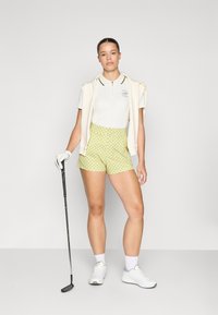 Bela polo majica s ovratnikom, zeleni detajli in logo; zeleni šortji s potiskom; lahek pulover, ovit okoli ramen; bele tenisice; golf palica.
