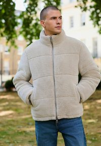 Jachetă din fleece bej, cu guler înalt, fermoar în față și buzunare laterale. Prezintă o textură matlasată cu cusături orizontale. Pur și simplu cu blugi albaștri.