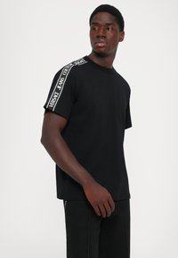 Άνδρας που φορά μαύρη μπλούζα με κείμενο "Versace Jeans Couture" στις ρίγες στους ώμους, στέκεται μπροστά σε λευκό φόντο.