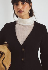 Must mustrip cardigan V-kaelusega, millel on kaks pruuni nuppu. Kandmine kerge ribaga kõrge kaelusega särk. Beez suède kott hoitakse kaenla all.