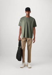 Oliivinvihreä puuvillainen t-paita, jossa kirjailtu logo, beige kapenevat housut, musta tikattu olkalaukku ja valkoiset lenkkarit. Mies seisoo suorassa ja käyttää mustaa lippalakkia.