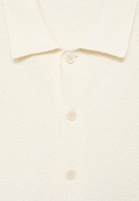 Polo shirt bianco in tessuto testurizzato con colletto piatto e cinque bottoni lungo la parte frontale, caratterizzato da un motivo sottile. Aspetto in tessuto morbido.