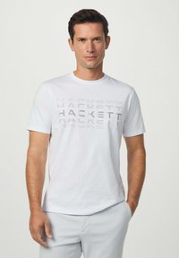 Bílý tričko s vyvýšeným šedým nápisem "HACKETT" na prsou, krátkými rukávy a kulatým výstřihem. Hladká bavlněná tkanina.