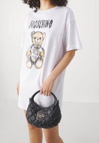 Kobieta w białej sukience T-shirt Moschino trzyma czarną pikowaną torebkę Love Moschino z uchwytami na tle jednolitym.