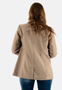 Cappotto in misto lana beige con spalle strutturate, cuciture sagomate e spacco sul retro. Abbinato a jeans in denim blu. Texture morbida, vestibilità sartoriale.