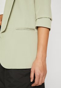 Světlý zelený blejzr z hladké látky s jedním náprsním kapsou a ohrnutými rukávy, doplněný černými kalhotami.