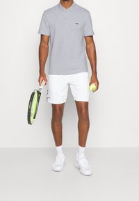 Szara koszulka polo z kołnierzykiem, białe szorty, trzymająca rakietę tenisową i żółtą piłkę tenisową. Strój uzupełniają białe buty sportowe.