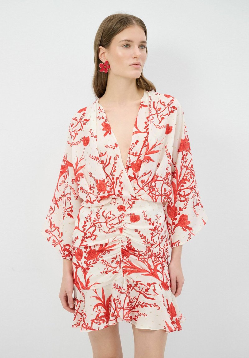 Een witte jurk met een bloemenpatroon en rode ontwerpen, voorzien van een diepe V-hals, kimono-mouwen en een gerimpelde taille. Glad materiaal met een geschulpte zoom.
