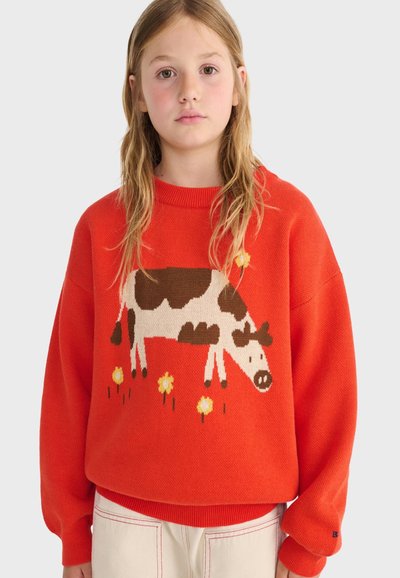 Jeune personne portant un pull orange avec une vache marron et blanche et des fleurs jaunes tricotées à l'avant.