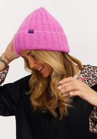 Ružová pletená čiapka z mäkkého materiálu, so žíhanou textúrou a malou značkou na prednej strane. Zúžená na vrchu s preklopeným lemom.