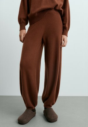 Partie inférieure du corps d'une personne portant un pantalon en maille marron ample, un pull assorti et des chaussures plates marron foncé, debout sur un sol gris.