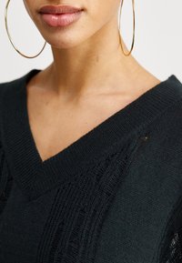 Svetr s výstřihem do V v tmavě zelené barvě, pletený z látky s texturovaným vzorem a otvorovými detaily podél výstřihu.