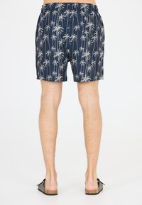 Marineblaue Bade shorts mit einem hellen Palmbaum-Muster, elastischem Bund und einem lockeren Schnitt, kombiniert mit dunklen Sandalen und barfuß.
