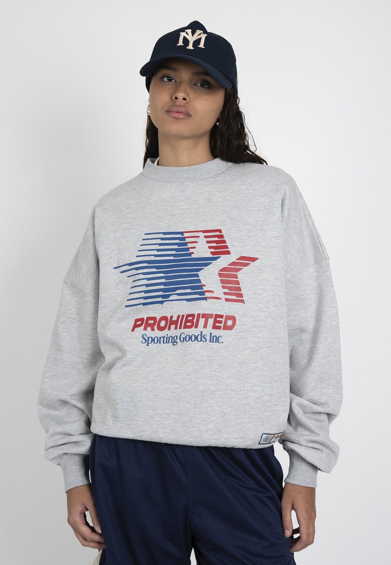 Jeune femme portant une casquette bleu marine et un sweat-shirt gris avec une étoile rayée rouge et bleue et le texte "PROHIBITED Sporting Goods Inc.", debout sur fond blanc.