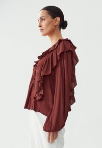 Maroon blouse featuring sheer ruffled sleeves and a button front. The fabric has a soft texture with a relaxed fit and loose silhouette.