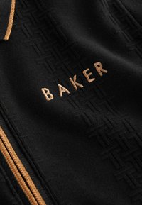 Sorte stofjakke med lynlås, der har et struktureret mønster og broderet "BAKER" i lys brun. Fremhævende detaljer inkluderer en kontrastfarvet lynlås.