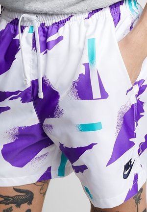 Witte shorts met paarse en turquoise abstracte patronen, een witte trekkoord, Nike-logo op het been en een gedeeltelijk zichtbare getatoeëerde been.