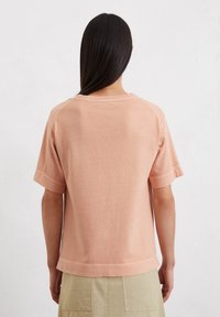 Korte mouwen, perzikkleurig shirt van zachte katoen. Heeft een ontspannen pasvorm, ronde halslijn en subtiele logodetail bij de achterkant van de hals.