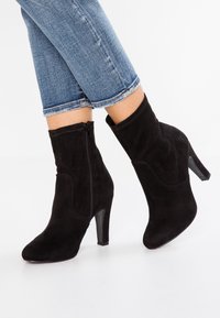Bottines en daim noir avec talon haut, dotées d'une fermeture éclair latérale, d'une texture lisse et d'une pointe effilée. Portées avec un jean bleu retroussé.