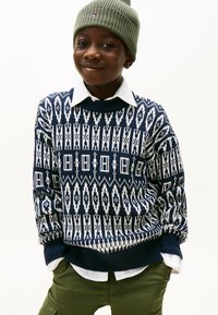 Navy- und weiß gemusterter Strickpullover mit Rundhalsausschnitt, über einem weißen Hemd mit Kragen getragen, kombiniert mit olivgrünen Hosen und einer grauen Mütze.