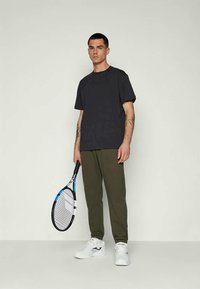 T-shirt nera in cotone abbinata a pantaloni jogger verde oliva, scarpe sportive bianche e in mano una racchetta da tennis con un design blu e nero.