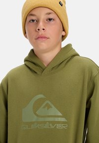 Oliivinvihreä huppari, jossa on suuri vaaleanvihreä Quiksilver-logo. Mallilla on päällään karkea keltainen pipo. Myös materiaaliltaan sileää kangasta, rento muotoilu.