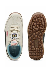 Baskets Puma avec une tige en toile crème, accents noirs, semelle intérieure bleue, semelle en caoutchouc dotée de petites grips circulaires et une étiquette en forme de personnage unique.