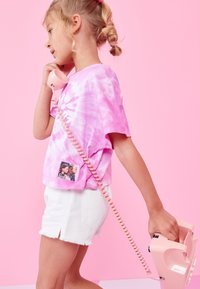 Niña joven con el cabello trenzado, vistiendo una camiseta rosa con efecto tie-dye y pantalones cortos blancos, sosteniendo y hablando por un teléfono rosa de disco.