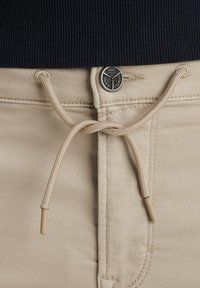 Beige bomuldsbukser har en snøre-lukning med plastiksnaps, en metalknap med branding og en ribbet marineblå taljeovenfor.
