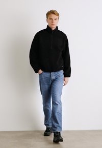 Zwarte fleece pullover met een halve rits, ontspannen pasvorm, gedragen met lichtblauwe spijkerbroek en zwarte sneakers. Eenvoudig ontwerp, geen zichtbare patronen.