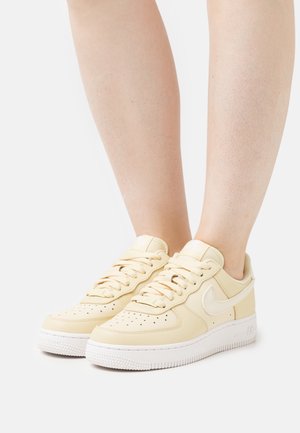 Une paire de baskets beige Nike Air Force 1 avec des semelles blanches, portées sur des jambes nues devant un fond blanc.
