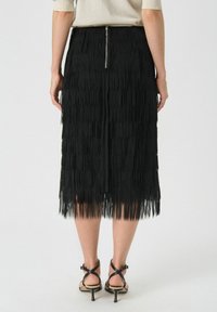Czarna spódnica z frędzlami o długości midi, wykonana z teksturowanego materiału z widocznym zamkiem błyskawicznym z tyłu, zestawiona z czarnymi sandałami na obcasie.