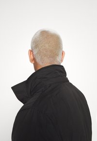 Man med kort grått hår som bär en svart jacka, sedd bakifrån mot en enfärgad vit bakgrund.