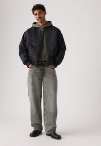 Μαύρο μπουφάν μπομπέρ με φερμουάρ μπροστά, φορεμένο πάνω από γκρι φούτερ με κουκούλα. Φαρδιά τζιν παντελόνια ανοιχτό γκρι και μαύρα δερμάτινα μποτάκια συμπληρώνουν το ντύσιμο.