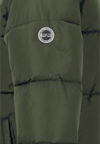 Veste coupe-vent verte avec une texture matelassée. Présente un écusson circulaire en argent au centre, affichant les détails de la marque.