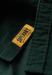 Groene corduroy stof met een zichtbare gele label waarop "ENGINEERED SUPERDRY HEAVY DUTY" in blauwe letters staat, wat een duurzaam ontwerp en textuur laat zien.
