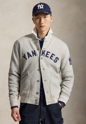 Fiatal férfi, aki szürke Yankees dzsekit, sötétkék logós sapkát, fehér inget felpöndörített gallérral, sötét farmernadrágot és órát visel, egy egyszínű háttér előtt áll.