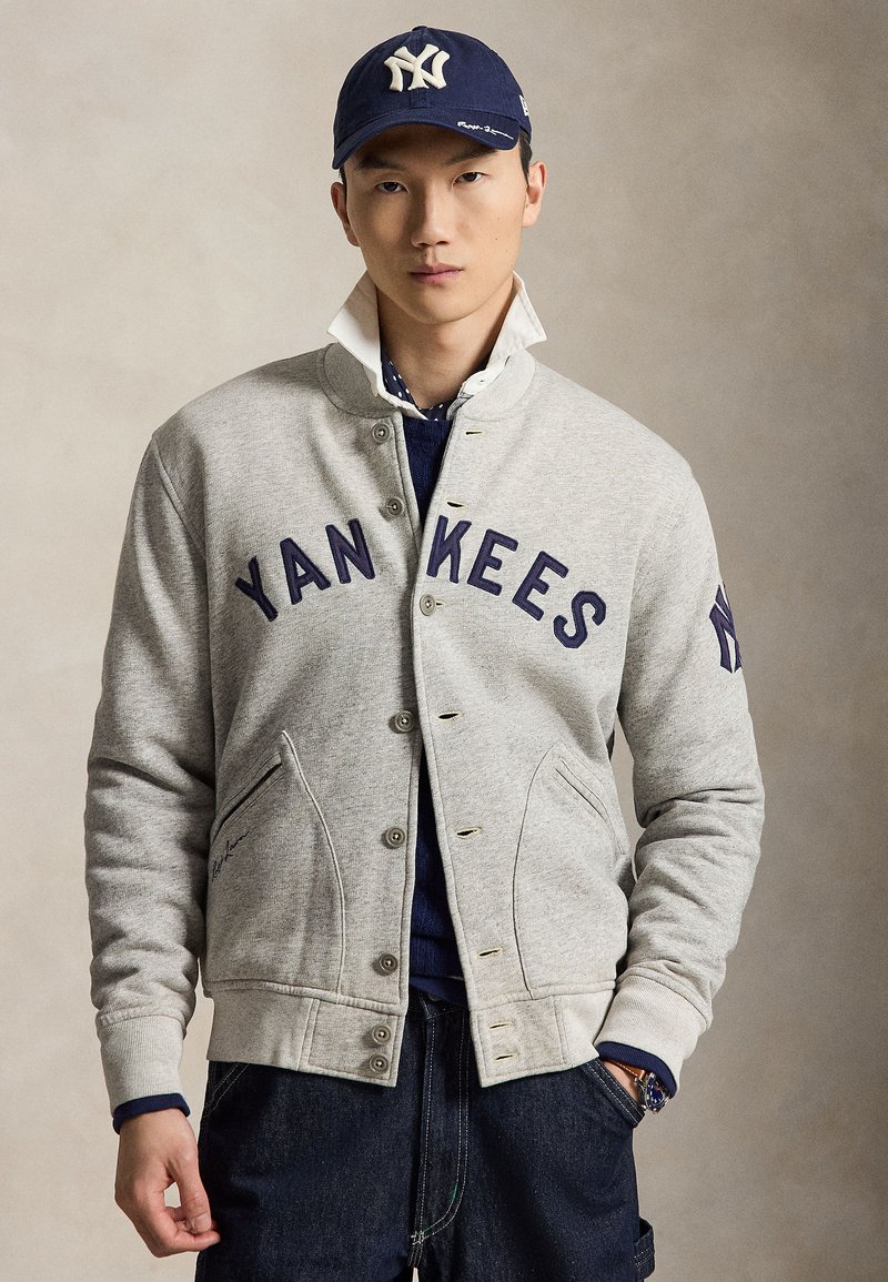 Jeune homme portant une veste grise des Yankees, une casquette bleu marine avec un logo, une chemise blanche au col relevé, un jean en denim foncé et une montre, debout devant un fond uni.