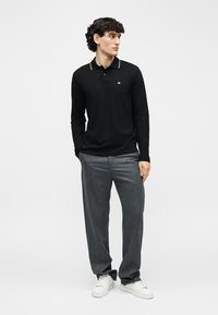 Vyras, vilkintis juodą ilgomis rankovėmis polo marškinėlį, pilkas kelnes ir baltus sportbačius, stovintis su rankomis kišenėse priešais paprastą baltą foną.