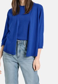 Blå cardigan med avslappnad passform, 3/4-långa ärmar, öppen framtill, ribbade manschetter och nederkant. Kombinerad med ljusblå högmidjade jeans.