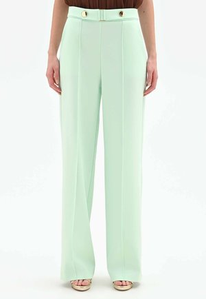Donna che indossa pantaloni a vita alta, gamba larga, verde menta con dettagli di bottoni dorati, abbinati a tacchi aperti beige.