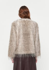 Faux fur jacket in beige with a textured, shaggy finish. Features a cropped silhouette and fringed edges. Worn over a brown skirt.