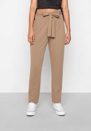 Frau trägt beige hoch taillierte, zulaufende Hose mit einem vorn zu einer Schleife gebundenen Stoffgürtel, kombiniert mit weißen Sneakern und einem schwarzen kurzen Oberteil.