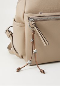 Beige strukturovaný kožený batoh se stříbrným zipem a hnědou koženou hvězdou jako přívěšek na klíče visící z jezdce přední kapsy.