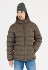 Brun puffjacka med dragkedja framtill, huva och elastiska ärmslut. Har en logotyp på bröstet och är tillsammans med blå jeans och en grön mössa.