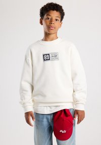 Sudadera blanca de cuello redondo, diseño holgado y parche de etiqueta azul marino. Gorra de béisbol roja sostenida en la mano con el logo. Jeans de mezclilla azul claro.