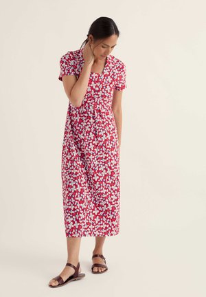 Nő piros midi ruhában, fehér és kék virágmintával, rövid ujjú, négyzet alakú dekoltázzsal és barna lapos szandállal, világos háttér előtt állva.